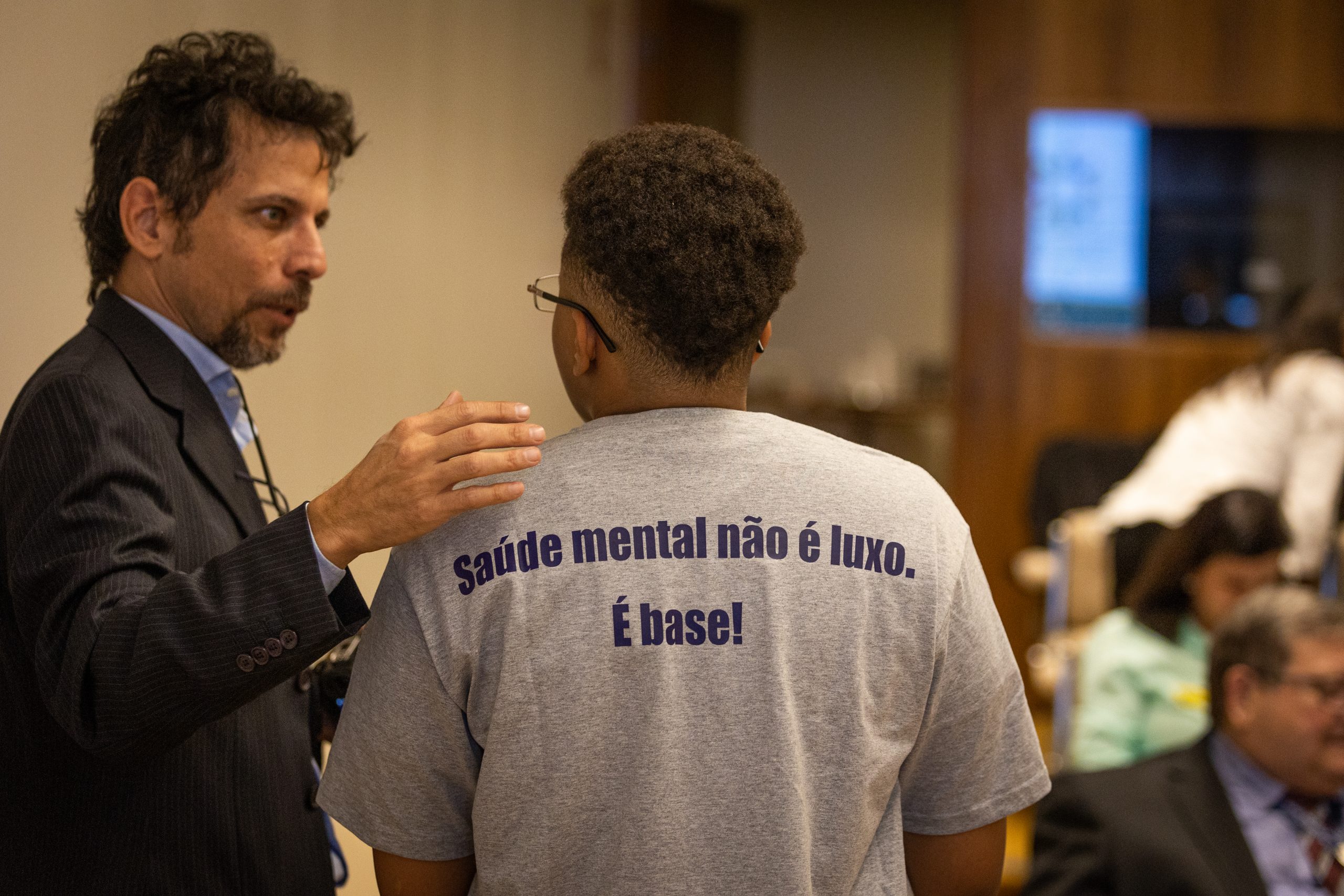 Homem branco de terno conversa com homem negro de camiseta cinza ao lado traz a frase “Saúde mental não é luxo. É base!”.