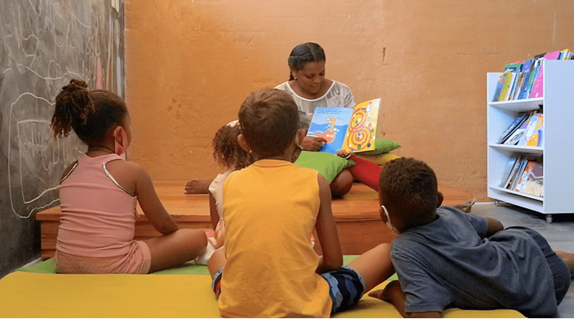 Mulher lê um livros para crianças, que estão de costas para a foto. Elas estão sentadas no chão.