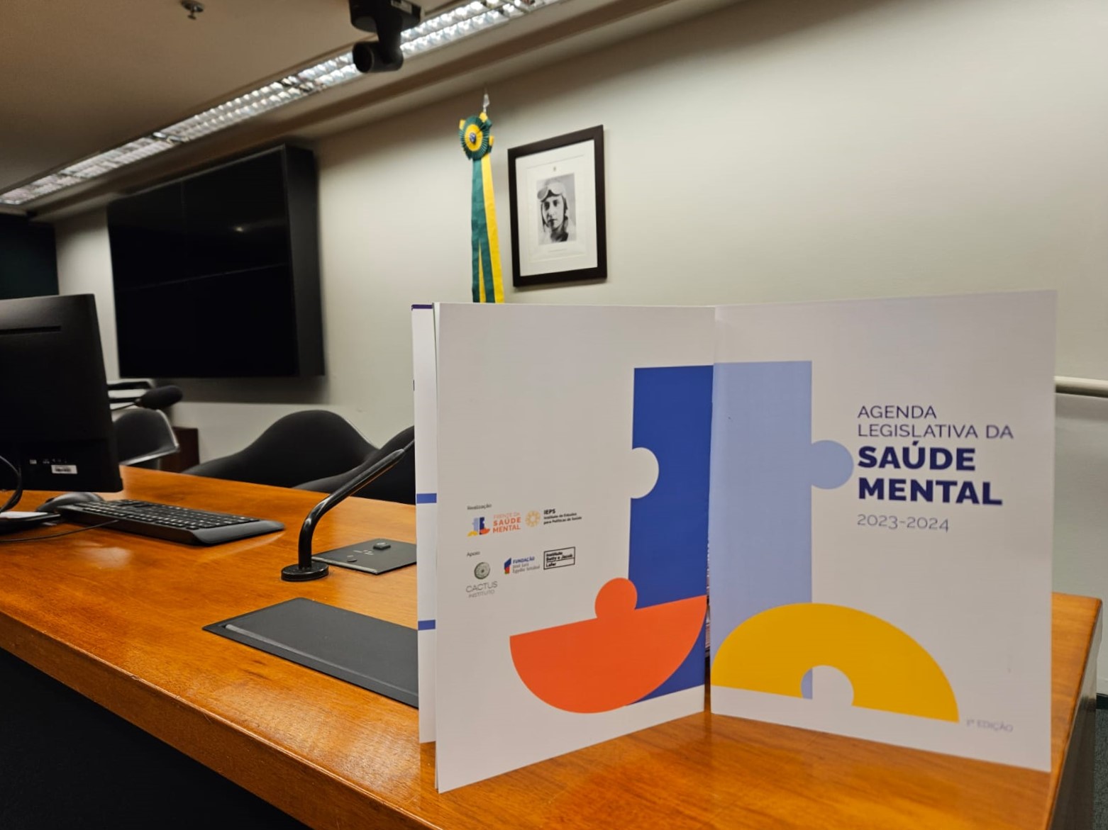 Livreto em mesa diretora com capa “Agenda Legislativa da Saúde Mental 2025-2026”, com ilustração colorida e ambiente de trabalho ao fundo, incluindo computador e bandeira do Brasil.