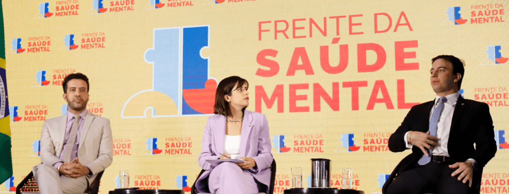 Painel do evento “Frente da Saúde Mental” com três participantes sentados em conversa, ao fundo há a estampa com o texto “Frente da Saúde Mental” e logo do evento.