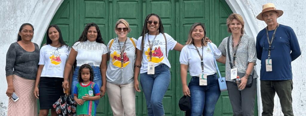 Grupo de pessoas em frente a uma grande porta verde, usando camisetas e credenciais de evento, com destaque para mulheres e uma criança. Foto externa em ambiente institucional.