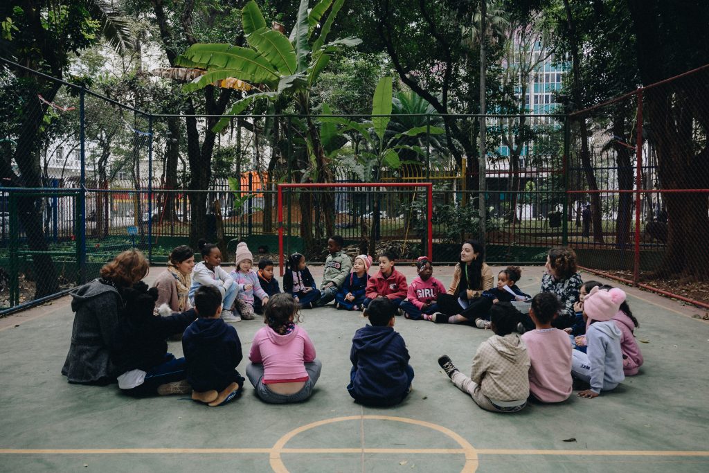Crianças sentadas em círculo no chão de uma área externa com piso quadriculado, cercas e árvores ao fundo, durante atividade em grupo no parque.