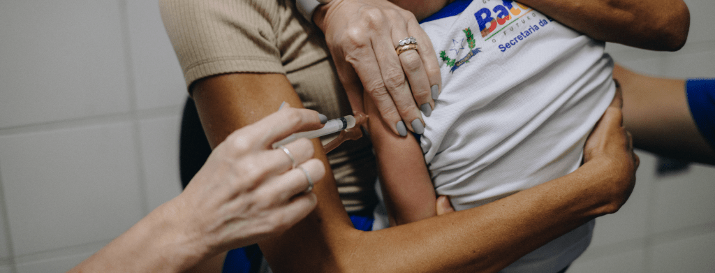 Equipe de saúde aplicando uma vacina em uma criança no braço, enquanto outras pessoas seguram e ajudam durante o procedimento de imunização em ambiente clínico.