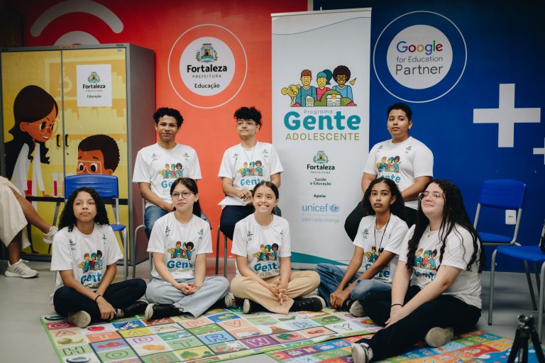 Equipe de jovens em evento educacional em sala decorada com logos “Fortaleza Educação”, “Gente Adolescente” e “Google for Education Partner”, sentados e em pé ao redor de um tapete colorido com letras.