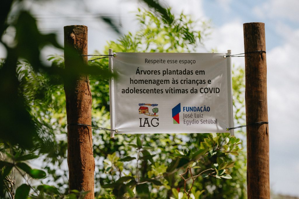 Banner em área externa preso entre dois troncos, com mensagem “Respeite esse espaço” e texto sobre árvores plantadas em homenagem a crianças e adolescentes vítimas da COVID, da Fundação IAG José Luiz Setúbal.